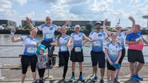 Gruppe von acht Menschen in Sportbekleidung steht an einem Ufergeländer vor einem Fluss oder Hafen; sie tragen Lauf- oder Radtrikots mit Nummernbändern und winken in die Kamera. Blau-weißer Himmel mit Wolken im Hintergrund, Boote und Gebäude am gegenüberliegenden Ufer sind sichtbar. Eine erwachsene Person hält ein Kleinkind auf dem Arm; alle wirken fröhlich und feierlich.