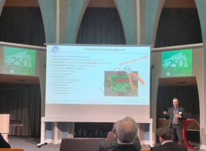 Konferenzsprecher steht rechts von einer großen Projektionsleinwand in einem Hörsaal mit gewölbten Deckenpaneelen. Auf der projizierten Folie sind Stichpunkte und ein Bild einer Leiterplatte zu sehen; auf den seitlichen Bildschirmen sind zwei kleinere grüne Folien mit '250' sichtbar. Im Vordergrund sitzen Zuschauer und blicken zum Sprecher.