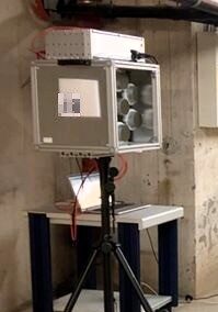 Technisches Messgerät in einem Kellerraum: eine offene graue Messbox auf einem schwarzen Stativ vor einer Betonwand, Kabel und Rohre im Hintergrund.