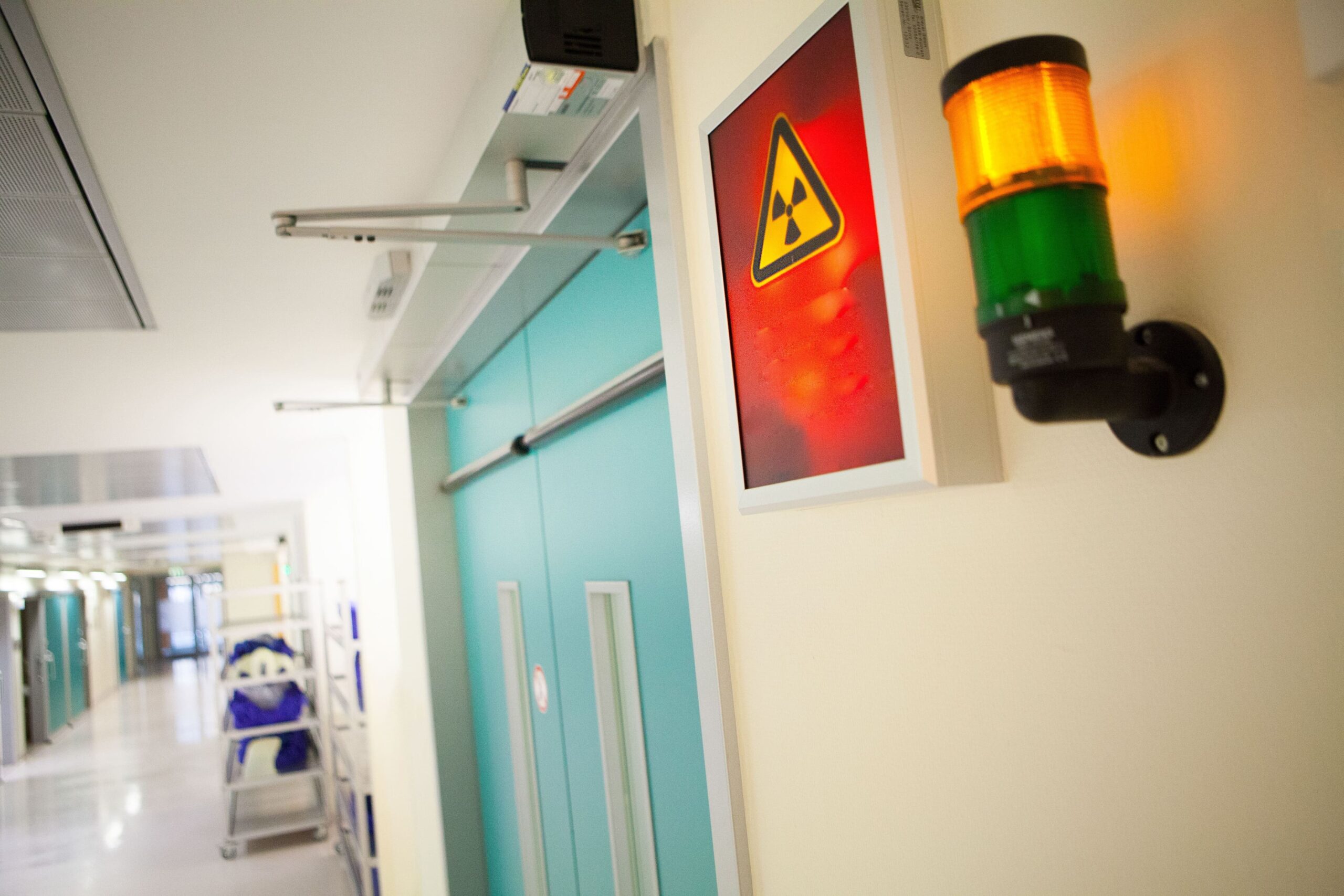 Flur in einer Radiologie: Deckenbeleuchtung und eine blinkende Warnleuchte mit gelb-orangefarbener Blitzsignallampe neben einem roten Warnschild mit Gefahrsymbol.