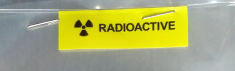 Durchsichtige Plastiktasche mit gelbem Warnaufkleber 'RADIOACTIVE' oben; darin eine runde, beige Scheibe mit aufgedrucktem blauen Logo