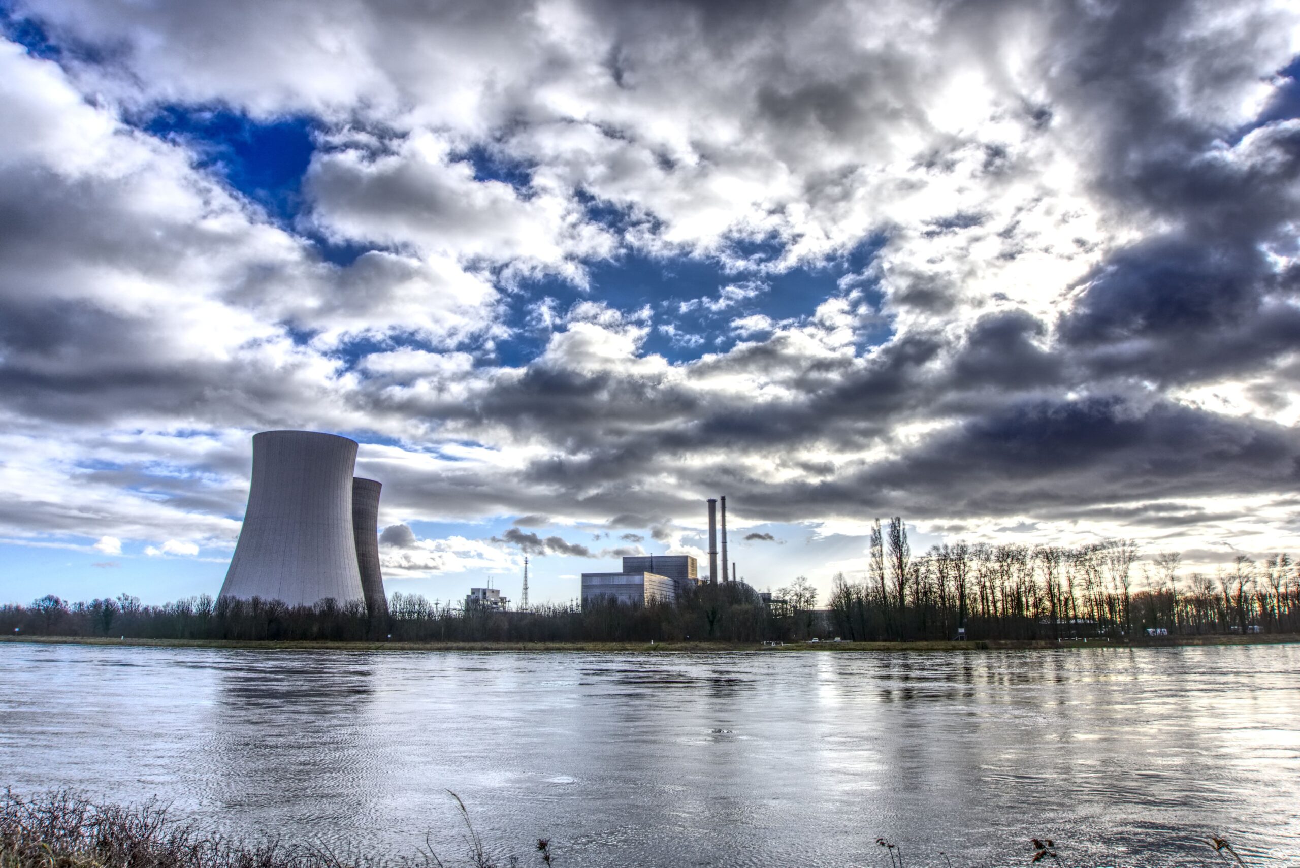 Stark bewölkter Himmel über einem Industriekomplex mit Kühlturm und Schornsteinen am Ufer eines breiten Flusses; flache Ufervegetation im Vordergrund.