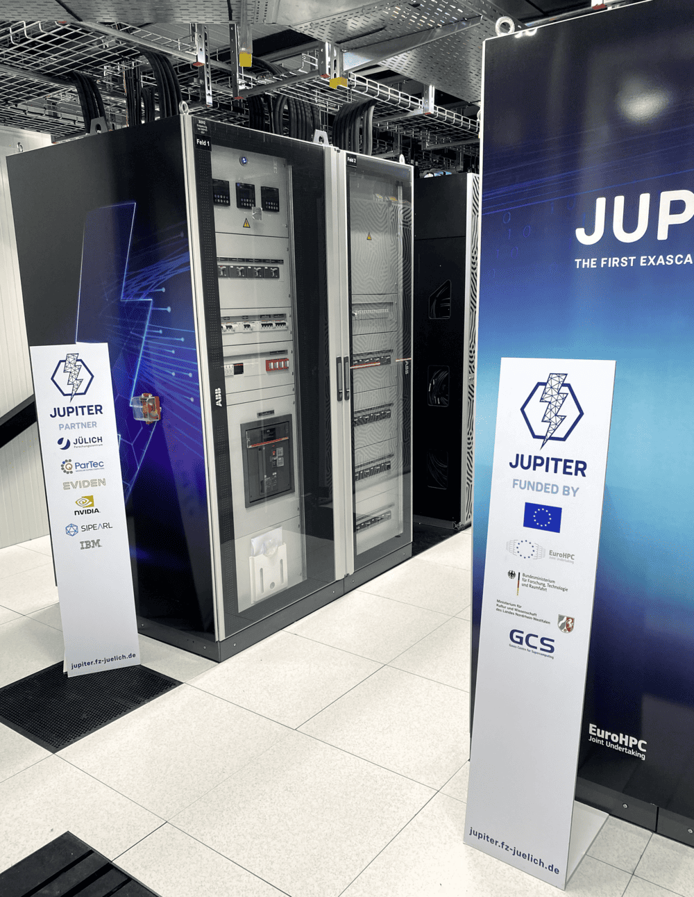 Blick auf einen Serverraum mit mehreren Schaltschränken/Racks und zwei Roll-Up-Werbeaufstellern mit der Aufschrift „JUPITER“ im Vordergrund.