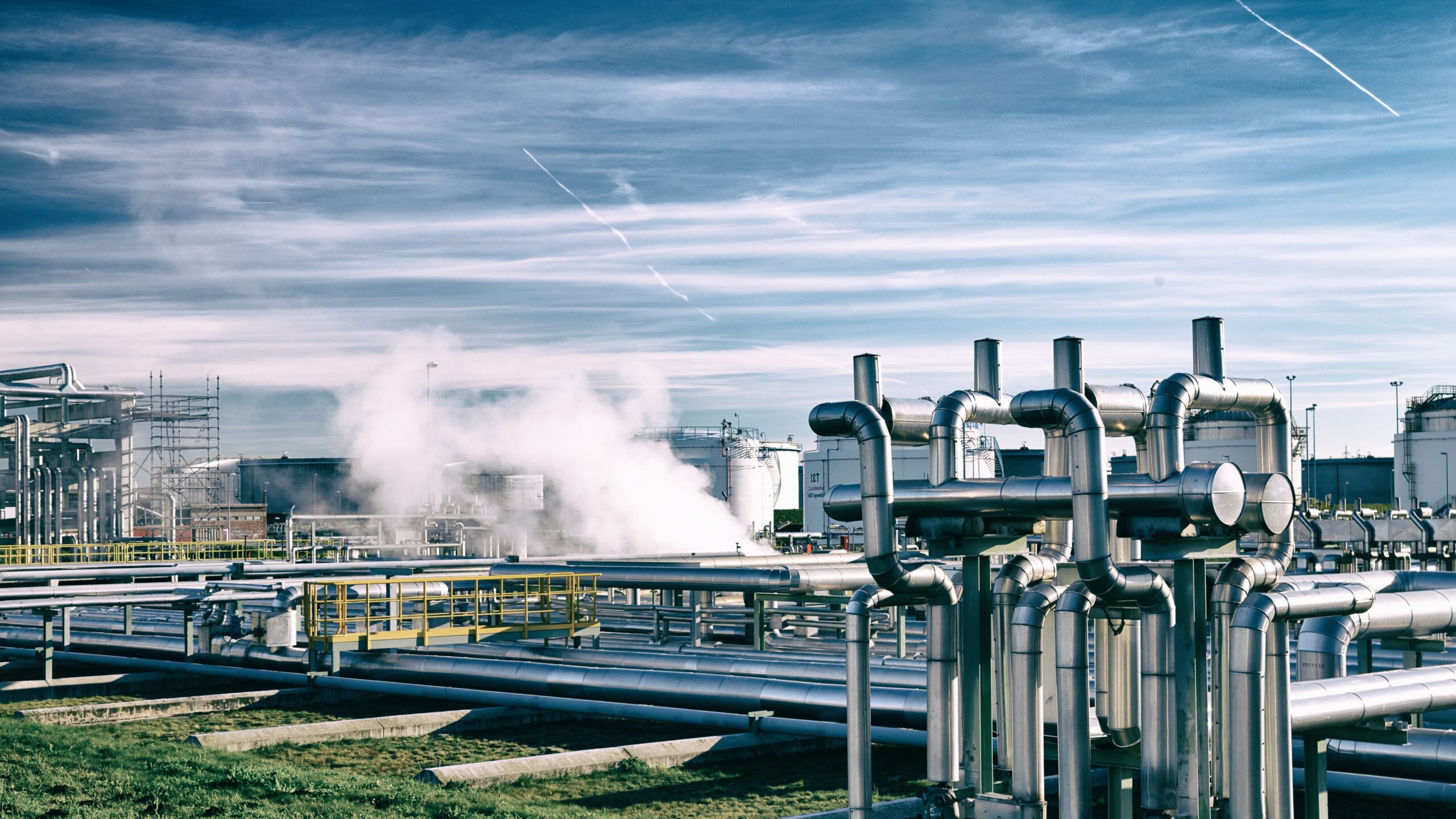 Industrielle Rohrleitungsanlage auf einer Energie- oder Chemieanlage bei Tageslicht: im Vordergrund mehrere große, verchromte Rohrleitungen und Ventile, im Hintergrund weitere Anlagen, Gitterplattformen und dampfende Leitungen; blauer Himmel mit dünnen Wolken.