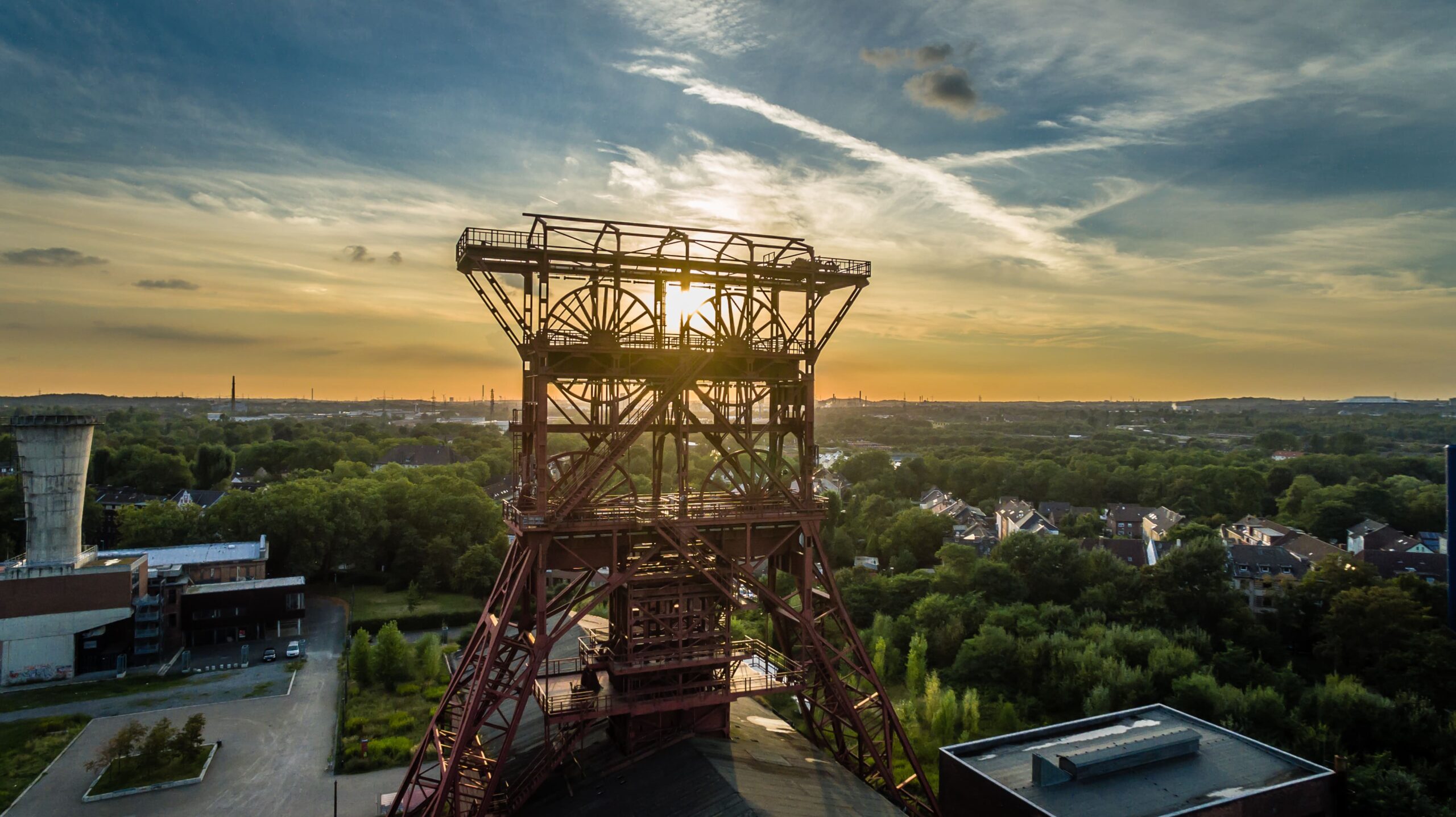 Ein großes, rostrot lackiertes Fördergerüst (Zeche) im Vordergrund, von oben gegen die untergehende Sonne fotografiert; die Sonne steht knapp über dem Horizont und hinterleuchtet die Stahlträger, im Hintergrund ein grüner Baumbestand, verstreute Gebäude und ein weiter Himmel mit Wolkenstreifen.