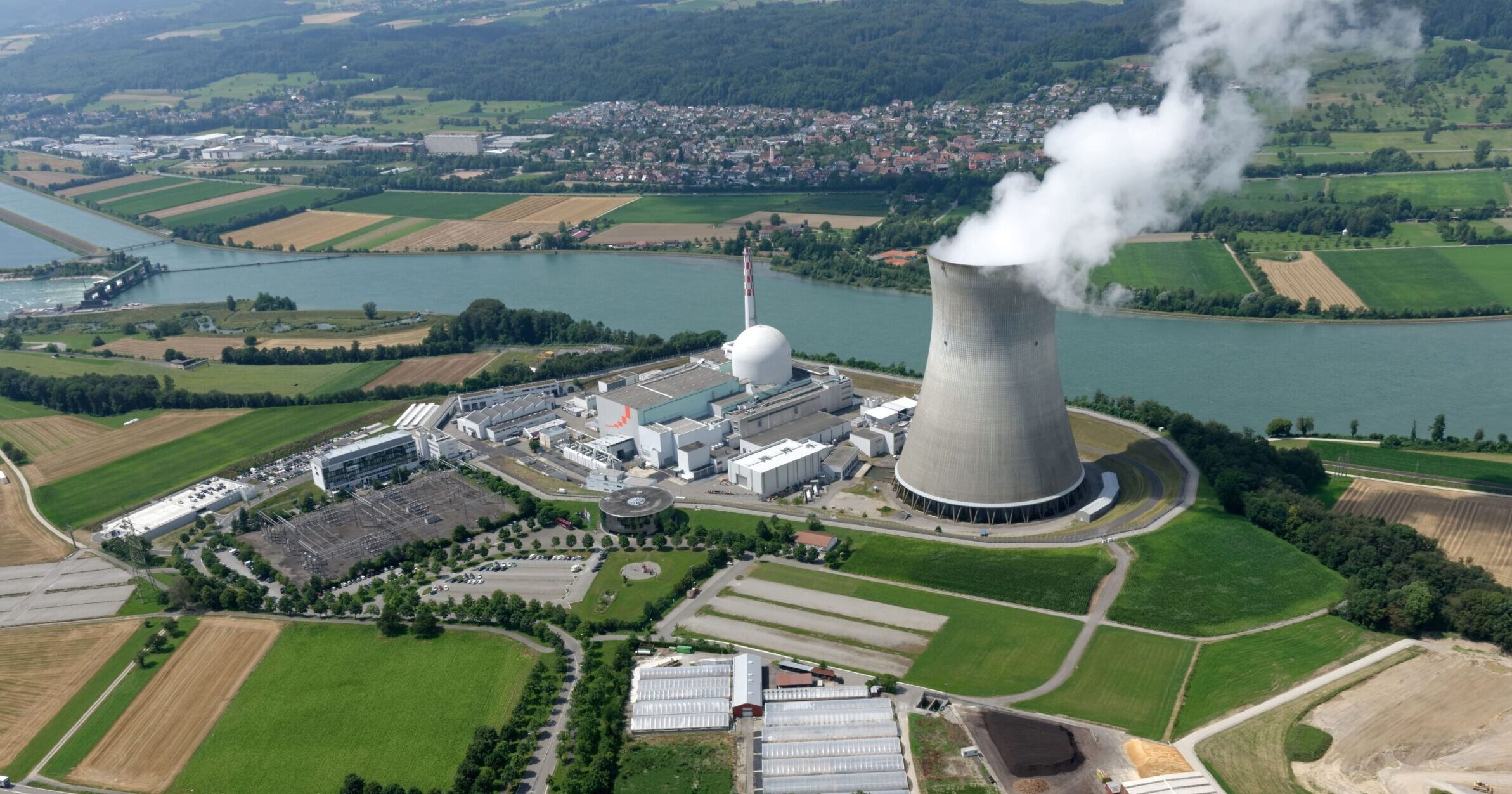 Luftaufnahme eines Kernkraftwerks an einem Fluss: im Vordergrund landwirtschaftliche Felder und Zufahrtsstraßen, auf dem Gelände mehrere Betriebsgebäude und ein markanter, rauchender Kühlturm sowie die kugelförmige Reaktorsicherheitsstruktur; Rauch oder Dampf steigt in einen klaren Himmel, Fluss und bewaldete Hügel im Hintergrund.