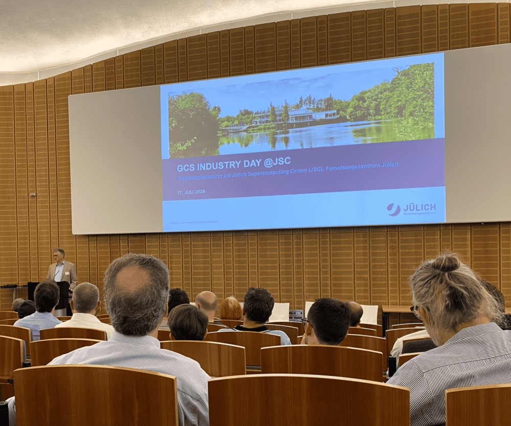 Vortragssaal mit Reihen von Holzsitzen, hinten mehrere Zuschauer; vorne links steht ein Redner am Pult, auf der großen Leinwand läuft eine Präsentationsfolie mit Bild eines Sees und Text „GCS INDUSTRY DAY @JSC“ sowie Logo „JÜLICH“; Raum hat gewölbte Decke und wandhohe, hellbraune Holzpaneele.