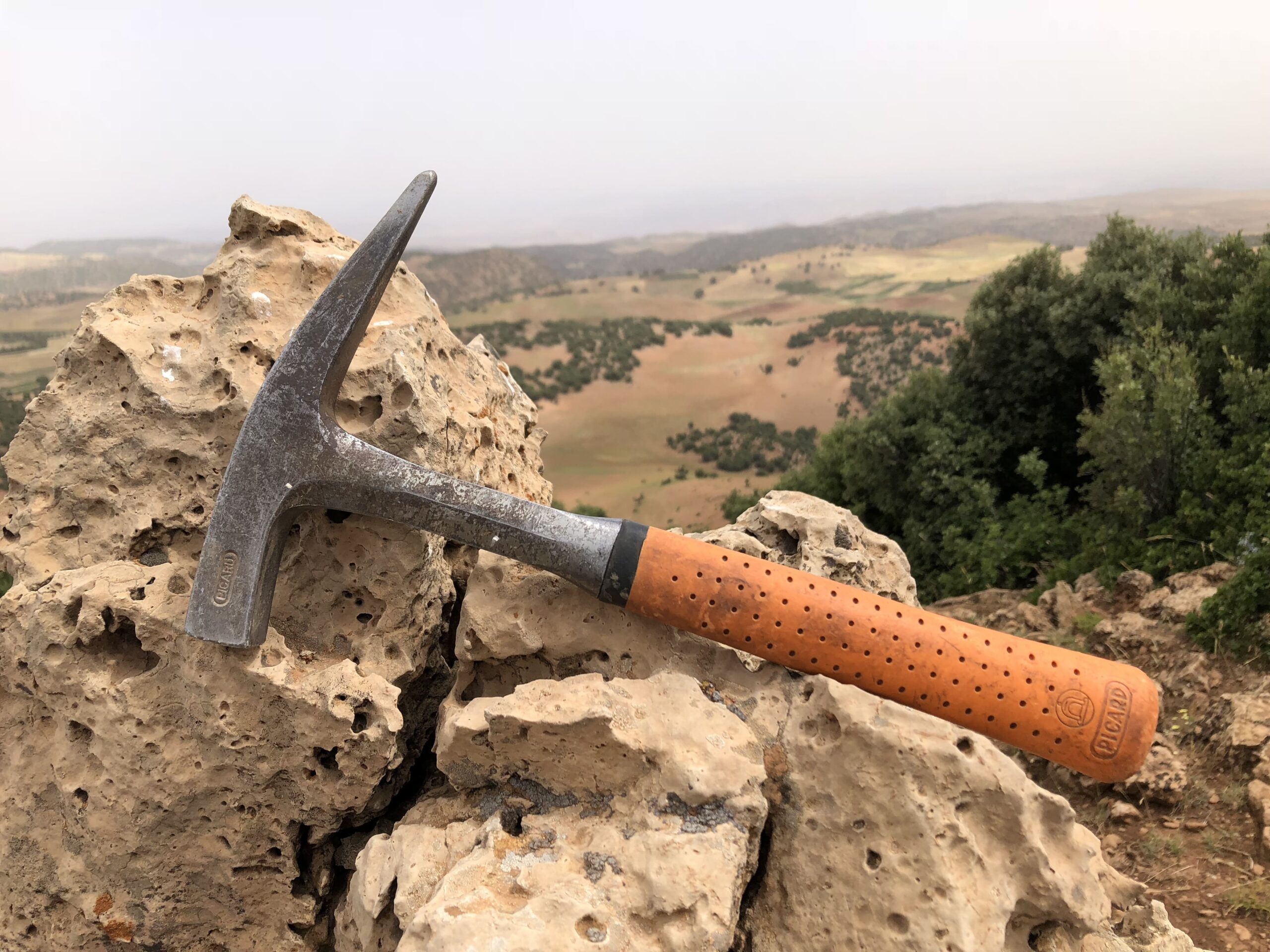 Geologischer Hammer mit orangefarbenem Griff liegt auf porösem Felsvorsprung; im Hintergrund hügelige Landschaft und vereinzelte Bäume unter hellem Himmel.
