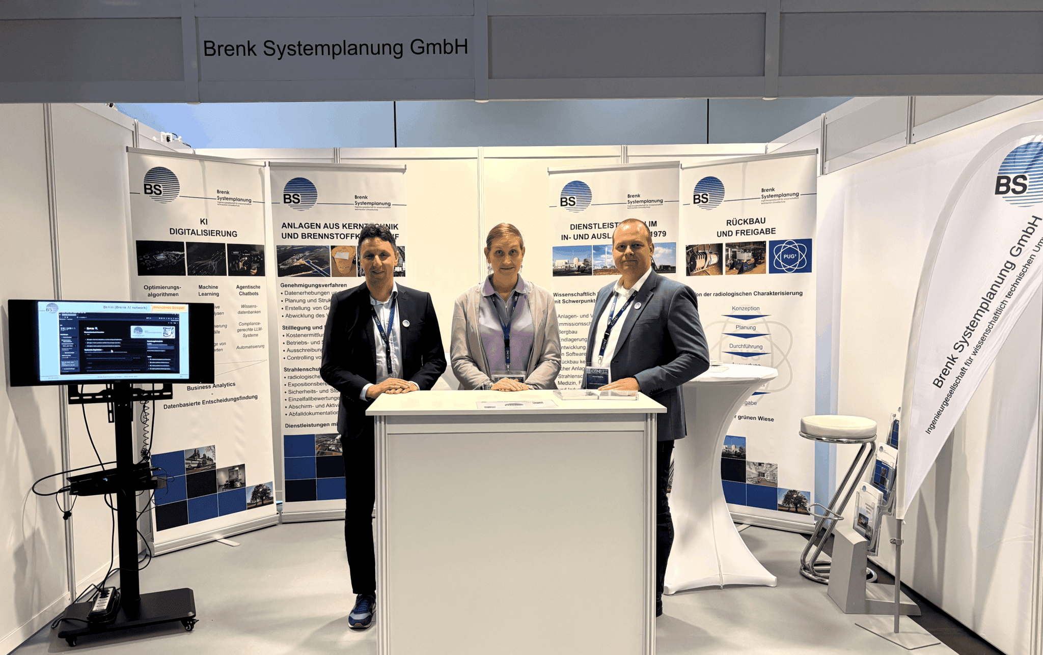 Drei Personen stehen an einem Messestand mit Informationswänden und einem zentralen Empfangstisch; links ein Bildschirm auf einem Ständer, rechts eine Stehtisch‑Ecke mit Hocker und einer gebogenen Messestand‑Grafik.