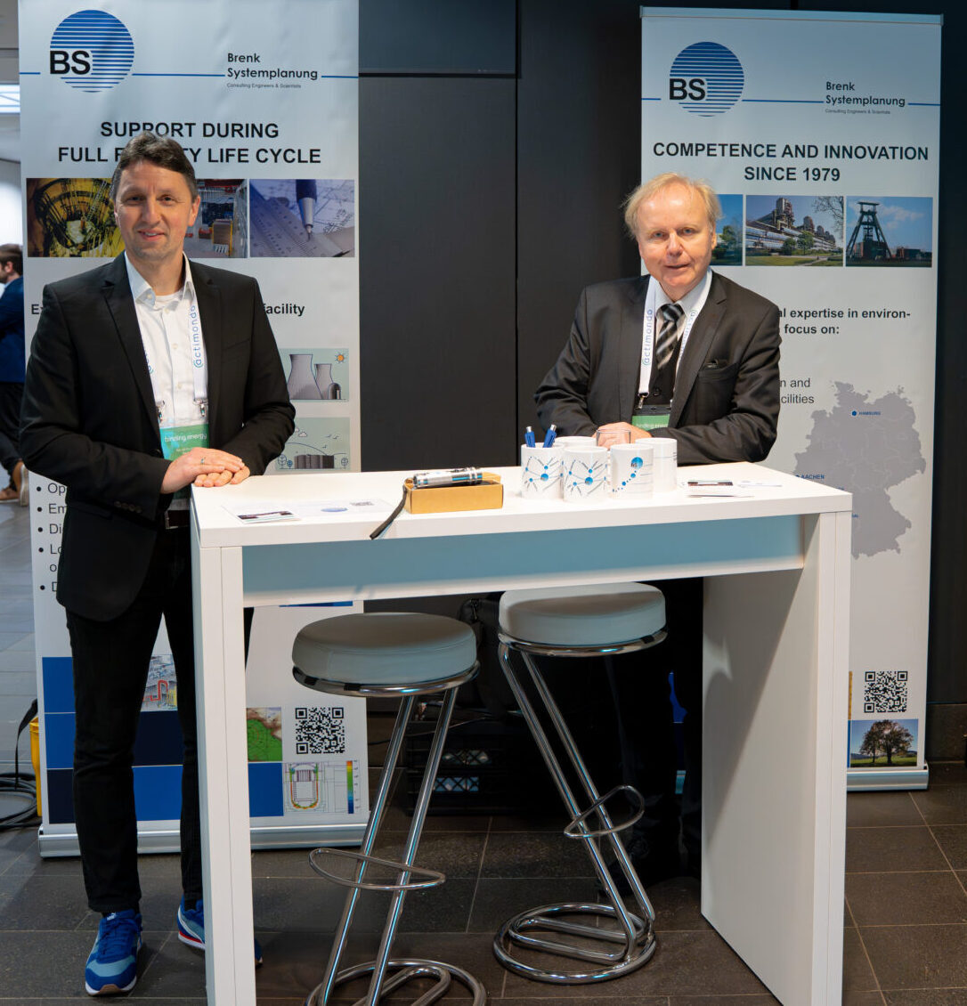 Foto auf einer Messe: zwei Männer stehen bzw. sitzen an einem kleinen Messestand mit Banner-Panels; im Vordergrund eine weiße Standtheke und ein Modell/Prüfaufbau mit Metallstützen sowie Informationsmaterial im Hintergrund.