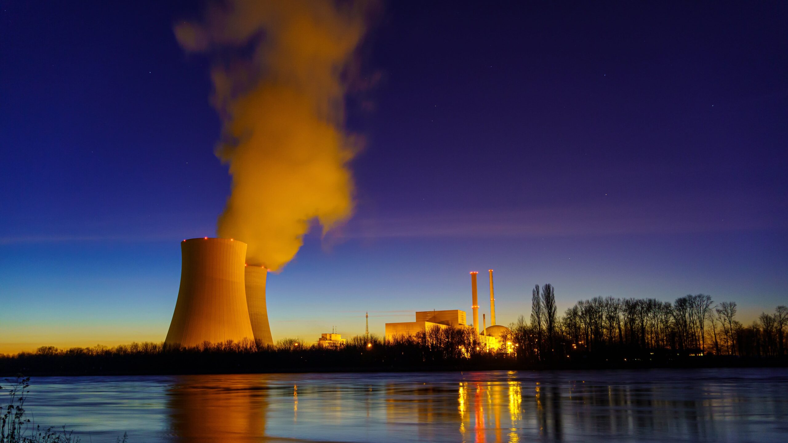 Kernkraftwerk am Wasser bei Dämmerung: im Vordergrund ein ruhiger Fluss mit Spiegelung der Lichter, im Hintergrund links ein grosser Kühlturm, aus dem dichter Dampf aufsteigt, rechts Gebäude und Schornsteine, am Horizont ein farbiger Sonnenuntergang und Silhouetten von Bäumen