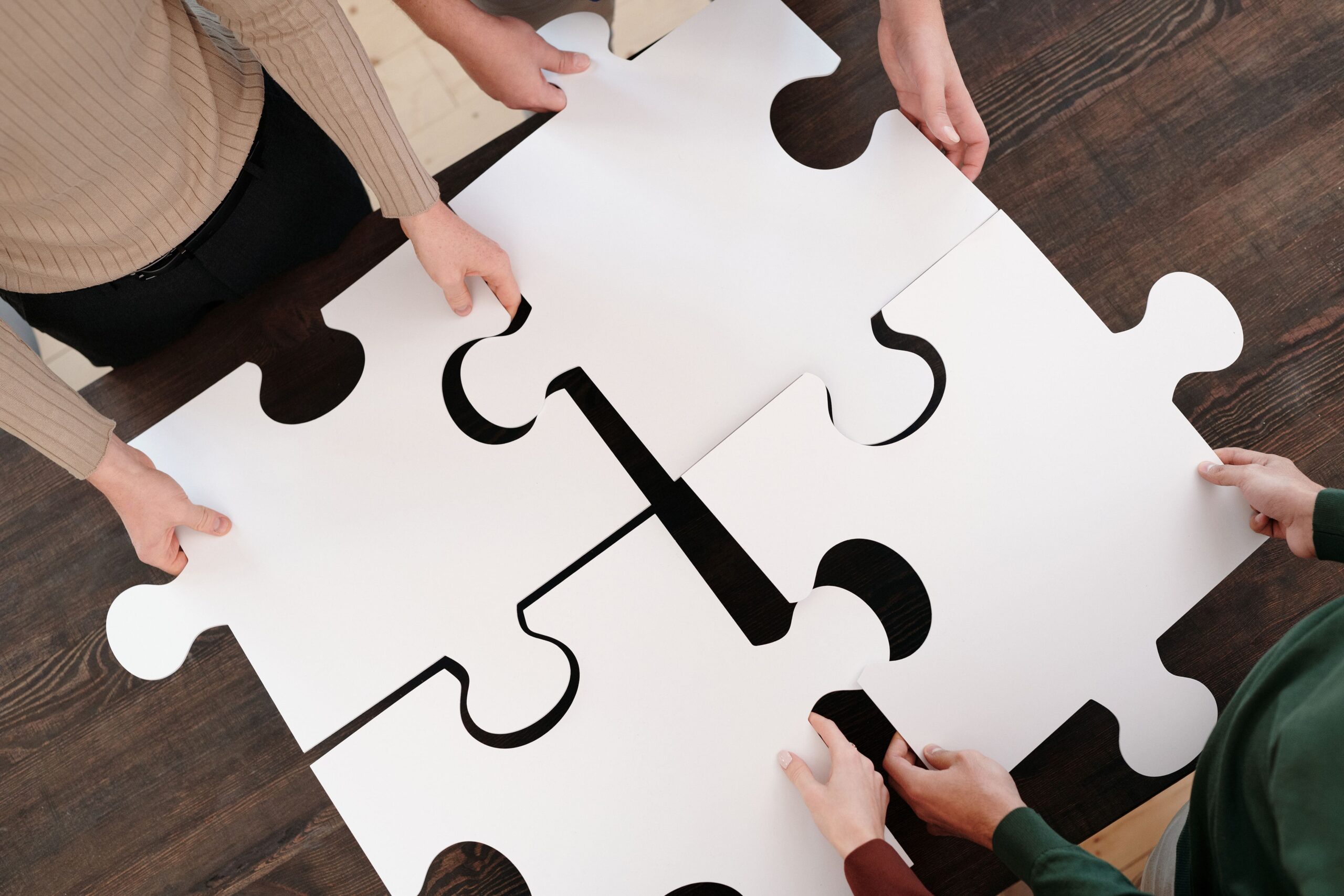 Vier Personen setzen von oben gesehen vier große, weiße Puzzleteile zusammen, die gemeinsam ein Quadrat bilden; man sieht nur die Hände und Teile der Arme auf einem dunklen Holzboden.