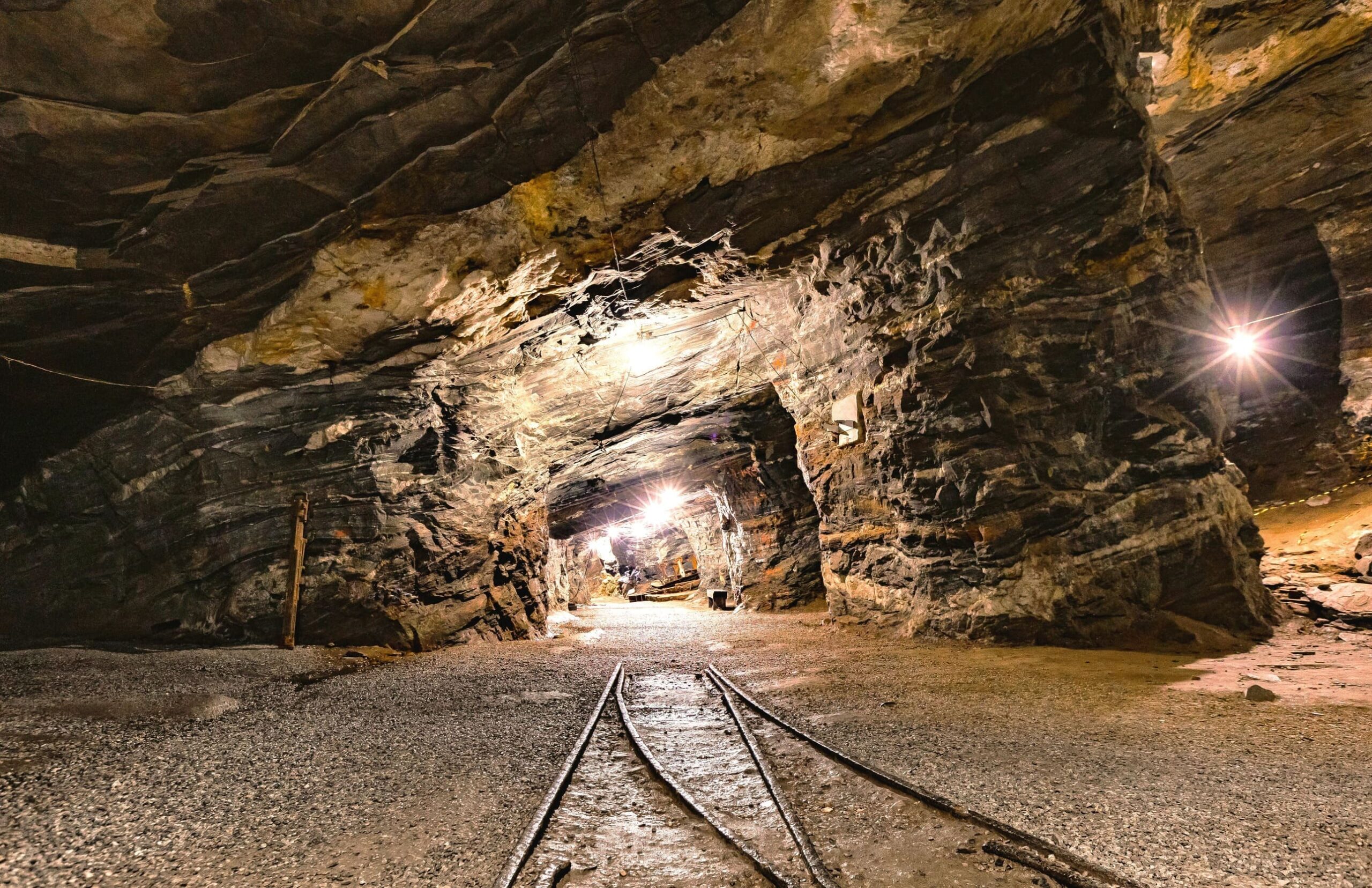Innenansicht eines bergwerksähnlichen Tunnels mit Gleisen in der Mitte, umgeben von steilen Felswänden; am Ende des Tunnels leuchtet ein Lichtschein.
