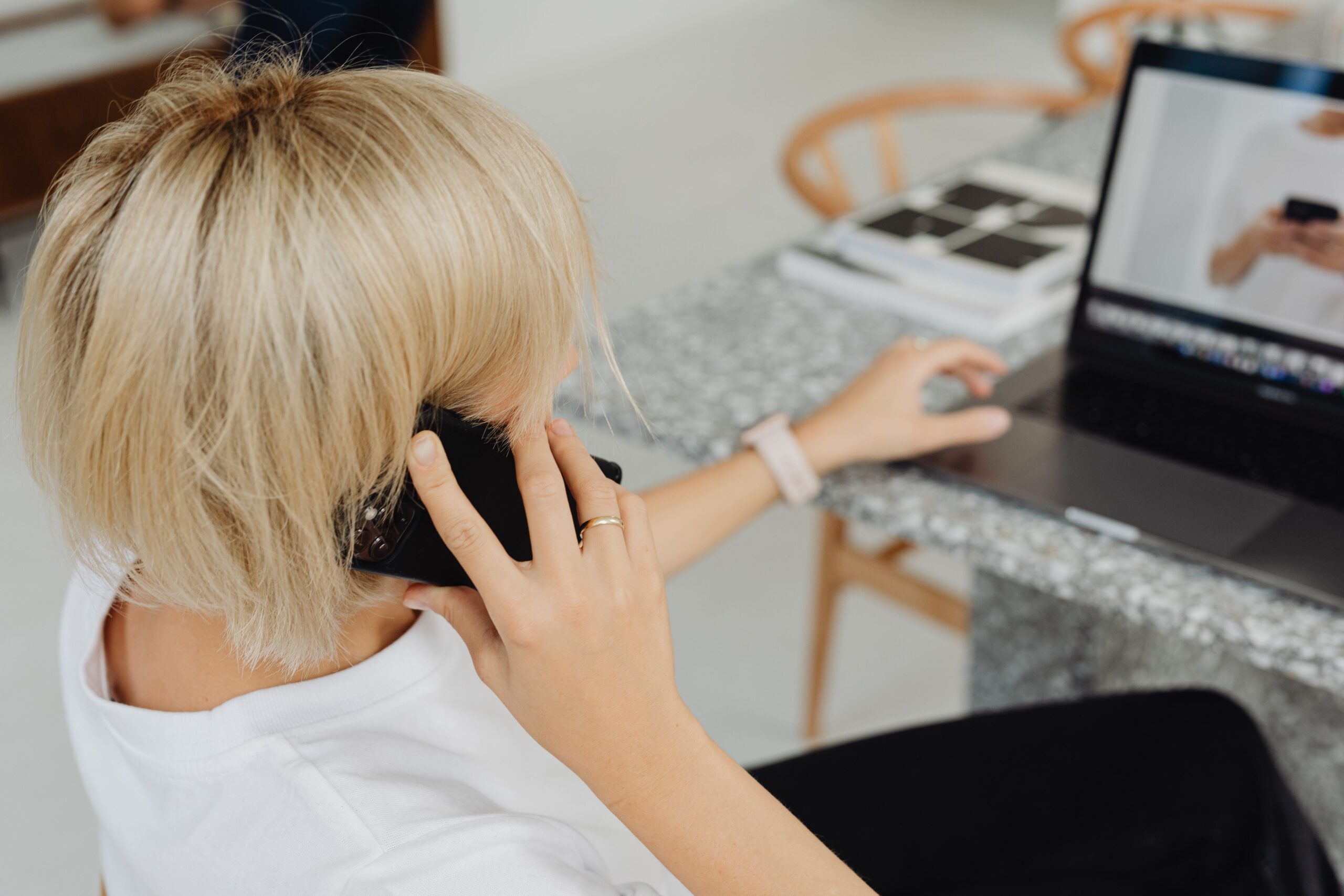 Eine Frau mit kurzem blondem Haar trägt ein weißes Oberteil und telefoniert im Sitzen, während vor ihr ein offener Laptop auf dem Tisch steht.