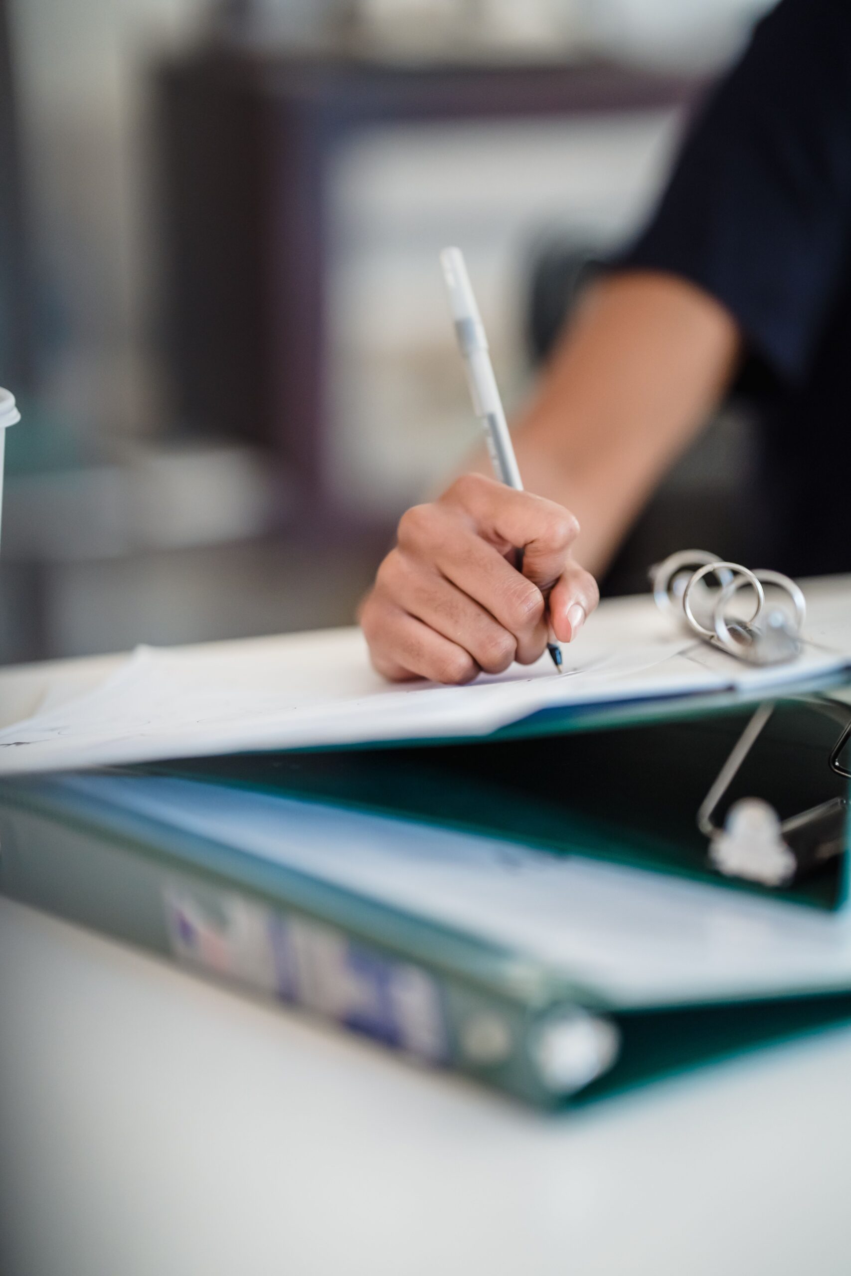 Person schreibt mit einem Stift auf Papier; im Vordergrund liegt ein aufgeschlagener Ordner mit Dokumenten und einem Ringmechanik-Einband.