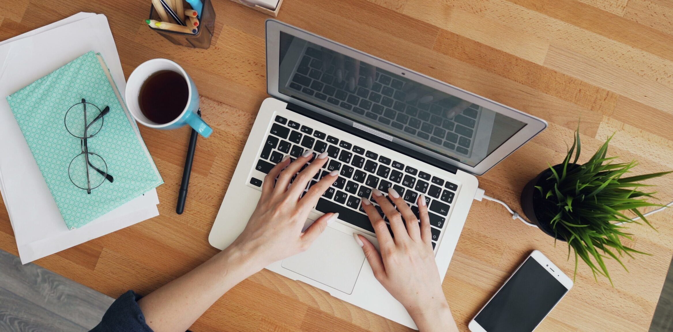 Eine Draufsicht auf einen Holzschreibtisch mit einer Person, die auf einem aufgeklappten Laptop tippt. Links liegen ein Notizbuch mit Brille, ein Stift und eine Tasse Kaffee; rechts ein Smartphone und eine kleine Topfpflanze. Auf dem Tisch stehen außerdem ein Stifthalter mit Büroklammern und Haftnotizen.