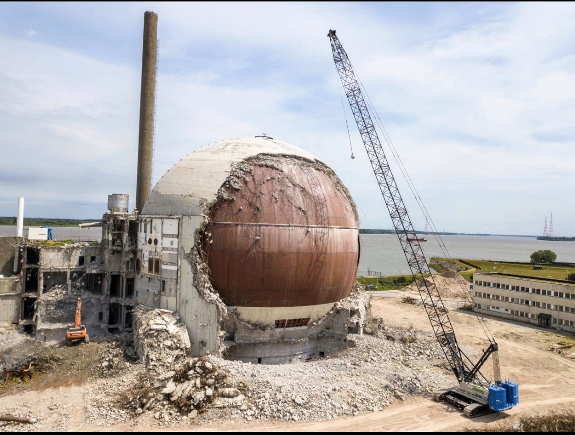 Blick auf eine große, zylindrische Beton- und Metallkugel mit geöffneter Bauöffnung, die auf einer Baustelle nahe dem Meer montiert wird, während ein Kran und weiteres Baugerät im Hintergrund stehen.