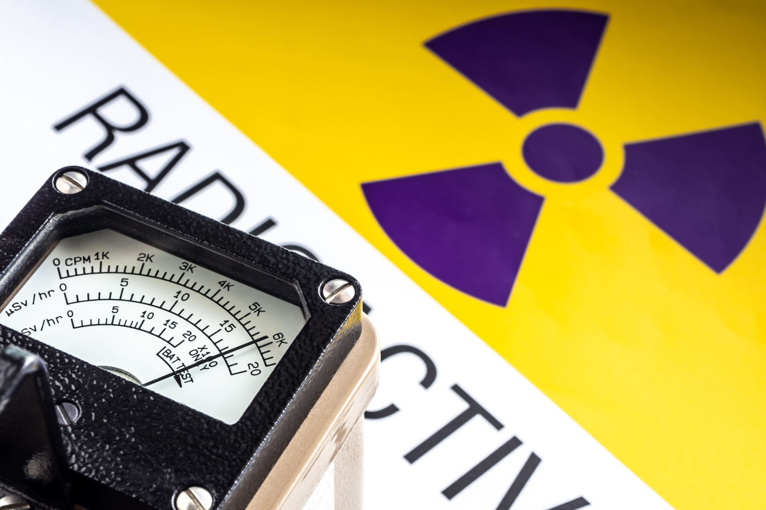 Nahaufnahme eines Messgeräts mit Skala und Sicherheitsaufschrift („RAD…“ im Hintergrund) vor einem gelb-violetten Hintergrund mit Strahlen-/Radioaktivitätswarnsymbol.